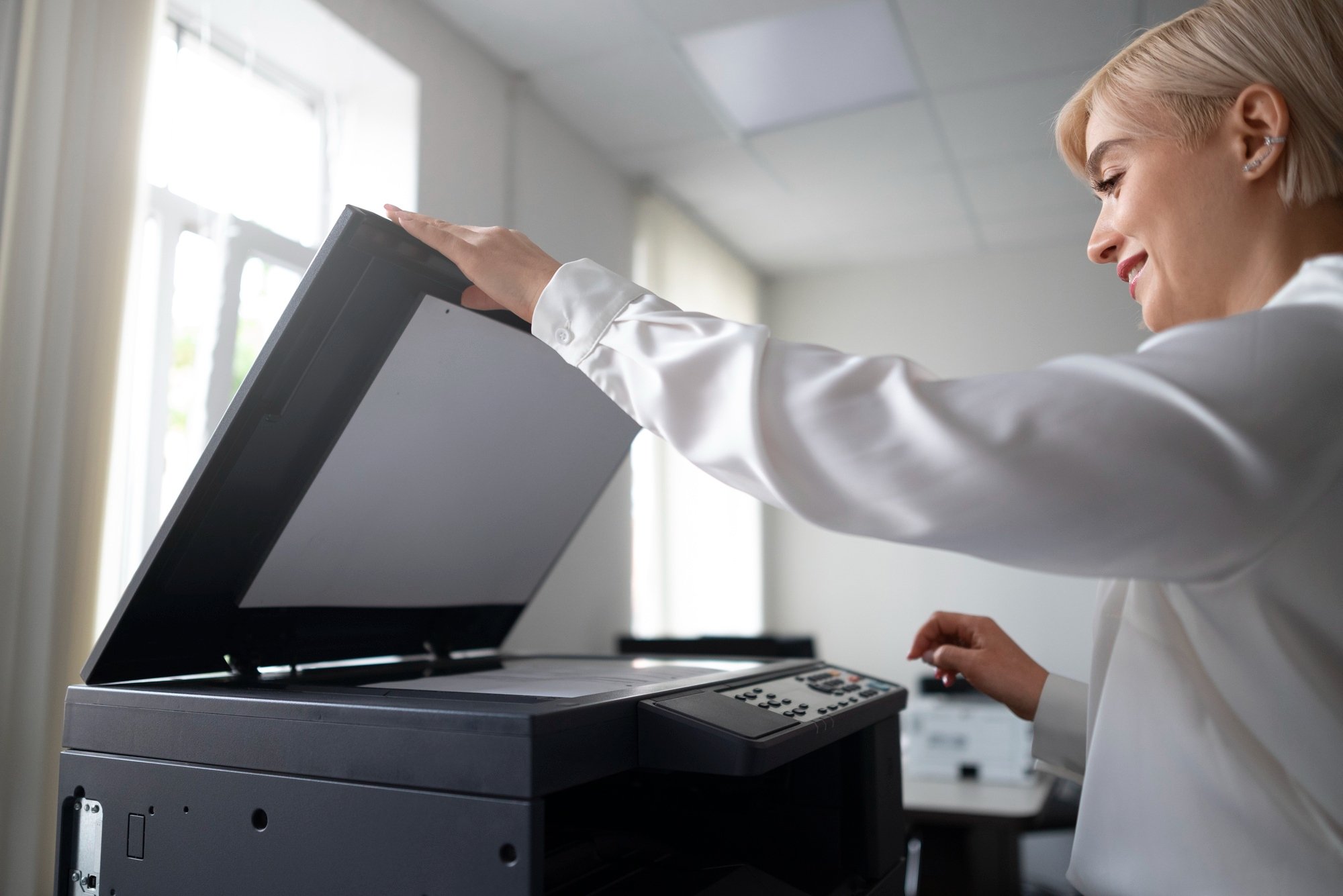A person in a white blouse smiles while operating a photocopier in a bright office.